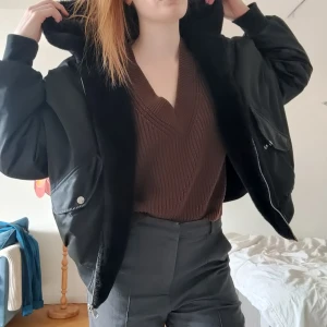 Svart vändbar bomberjacka - Svart bomberjacka med mjukt mysigt foder som är vändbar, så du får 2 jackor i 1! Jackan har dragkedja och tryckknappar på fickorna samt en ficka med dragkedja på ärmen. Stor och mysig luva. Perfekt för kyligare dagar. 