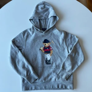 Grå hoodie från Ralph Lauren med Polo Bear - Säljer en grå hoodie från Ralph Lauren med en cool Polo Bear-tryck på framsidan. Tröjan har långa ärmar och en klassisk huva. Perfekt för en avslappnad stil. 🐻st. L för 14-16 år