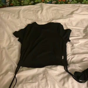 Svart topp från Shein - Säljer en svart topp från Shein med korta ärmar och knytband i sidorna. Perfekt för en stilren look. Toppen är tillverkad i ett mjukt material som känns skönt mot huden.