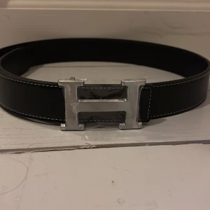 Svart bälte med silvrigt H spänne - Snyggt svart bälte med ett stort, silverfärgat H-format spänne. Bältet har vita sömmar längs kanterna som ger en stilren kontrast. Perfekt för att ge en elegant touch till din outfit.