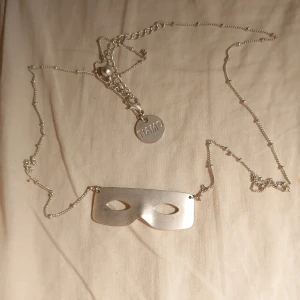 Silverfärgat halsband med maskberlock - Unikt silverfärgat halsband med en maskformad berlock. Kedjan är tunn och elegant, perfekt för att ge en subtil men stilfull touch till din outfit. Kedjan är längre än ditt standard halsband, så tänk på det! Får det nästan runt midjan. Det står TIAMO på den lilla platta cirkeln.