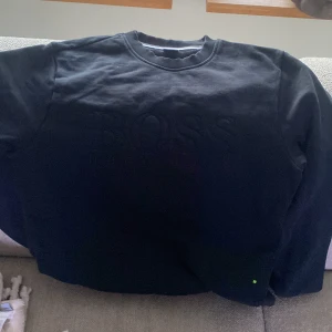 Svart sweatshirt från Hugo Boss - Säljer en stilren svart sweatshirt från Hugo Boss med diskret broderad logga på bröstet. Tröjan har en rund halsringning och ribbade muddar vid ärmslut och nederkant. Perfekt för en avslappnad och trendig look.