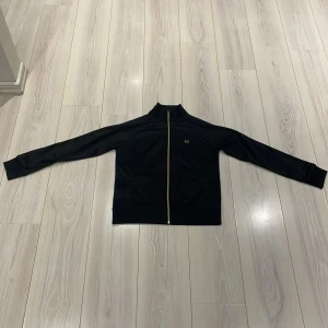 Svart tröja med zip från Fred Perry - Säljer en stilren svart zip up tröja från Fred Perry med gulddragkedja och broderad logga på bröstet. Jackan har långa ärmar och en klassisk passform. Nypris ligger på 1500kr, Perfekt för en sportig look eller casual vardagsstil.