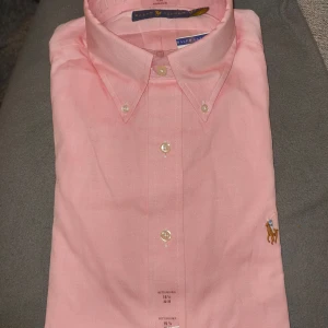 Oanvänd Ralph Lauren Skjorta - Oanvänd ny rosa skjorta från Ralph Lauren med klassisk krage och knappar framtill. Skjortan har en broderad logotyp på bröstet och är perfekt för en elegant look.