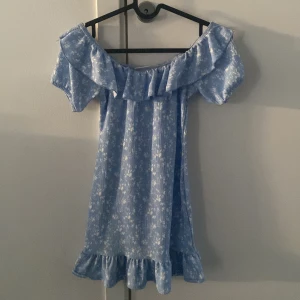 Blå offshoulder klänning från Gina Tricot - Söt blå offshoulder klänning från Gina Tricot med volangdetaljer och ett ljust blommönster. Perfekt för sommarens varma dagar. Klänningen har en luftig passform och är tillverkad i ett mjukt material.