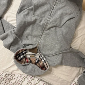 Grå huvtröja från Burberry - Säljer en stilren grå huvtröja från Burberry med dragkedja och klassiskt rutigt mönster på insidan av huvan. Tröjan har långa ärmar och en mjuk insida för extra komfort. Perfekt för en avslappnad och trendig look. Tagen ingår ej. Liten fläck på ärmen. Men inget man ser. 