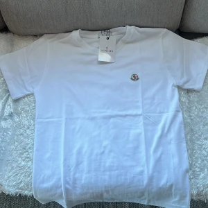 Vit t-shirt från Moncler - Säljer en stilren vit t-shirt från Moncler med korta ärmar och en liten broderad logotyp på bröstet. Perfekt för en avslappnad och snygg look. Passar bra till både jeans och shorts.