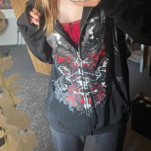Svart skull hoodie - Säljer en svart hoodie med dragkedja och stort tryck av dödskallar, röda rosor och vita detaljer på framsidan. Jackan har lång ärm och en avslappnad passform. Perfekt för dig som gillar alternativ stil 🖤