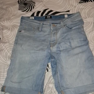 Ljusblå jeansshorts från Jack & Jones - Ljusblå jeansshorts från Jack & Jones med klassisk femficksdesign och uppvikta benslut. Shortsen har bälteshällor och knappgylf. Perfekta för varma dagar.
