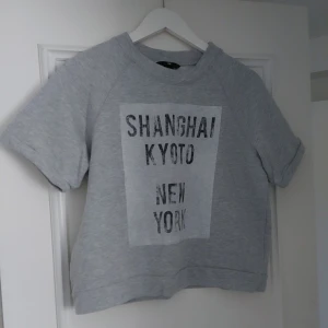 Grå t-shirt med tryck från H&M - Grå t-shirt från H&M med svart texttryck på bröstet: 'Shanghai Kyoto New York'. T-shirten har korta ärmar och en avslappnad passform. Perfekt för en cool och enkel stil.