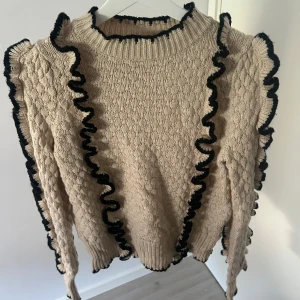 Beige stickad tröja med volang från Object - Säljer en beige stickad tröja från Object med svarta volangdetaljer längs ärmar och fram. Tröjan har lång ärm, ribbad hals och muddar. Perfekt för dig som vill ha något unikt och trendigt i garderoben!💗
