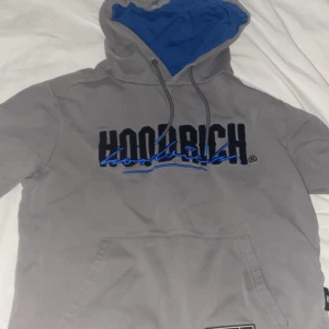 Grå hoodie från Hoodrich - Säljer en grå hoodie från Hoodrich med svart och blå logga broderad på bröstet. Tröjan har huva med dragsko och en stor magficka framtill. Perfekt för en avslappnad stil.