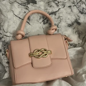 Ljusrosa handväska med gulddetaljer - Säljer en jättesöt ljusrosa handväska med snygga gulddetaljer och dekorativa knutar på handtaget. Väskan har flera fack inuti och stängs med både dragkedja och magnetlås. Perfekt storlek för det viktigaste och en stilren accessoar till din outfit. Köpt på Öland för runt 300 kr 