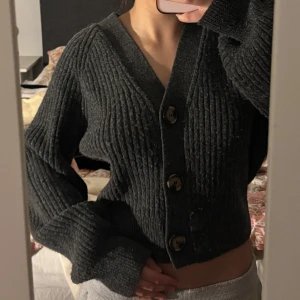 Mörkgrå croppad kofta med stora knappar - Säljer en mörkgrå, croppad kofta med stora knappar framtill. Tröjan är stickad med bred ribbad struktur och har långa ärmar samt djup v-ringning. Perfekt för lager-på-lager och en avslappnad stil.