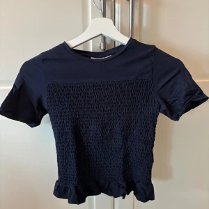 Mörkblå topp med smock från Gina Tricot - Säljer en mörkblå topp från Gina Tricot med smockad front och baksida samt volangkant nedtill. Toppen har korta ärmar och rund halsringning. Perfekt för en stilren och trendig look.