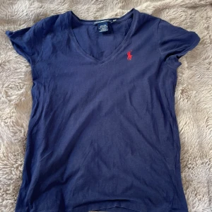 Mörkblå t-shirt från Ralph Lauren Sport - Mörkblå t-shirt från Ralph Lauren Sport med v-ringning och röd broderad logga på bröstet. Tillverkad i 100% pima bomull för en mjuk och skön känsla. Perfekt för sommaren!🥰 Har aldrig använt då jag bedömer den som ny💓. Den är äkta, skriv privat för fler bilder!🥰