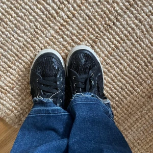 Svarta sneakers med spetsdetaljer - Säljer ett par svarta sneakers med snygga spetsdetaljer och mönstrad ovandel. Skorna har vita sulor och klassisk snörning. Perfekta för dig som vill ha en unik touch på dina vardagsskor.