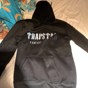 Svart hoodie med tryck från RATSART - Säljer en svart hoodie med stor ficka framtill och tryck i vitt och grått på bröstet där det står 'RATSART IT'S A SECRET'. Perfekt för en avslappnad och cool stil. Huva med dragsko och mjukt material.