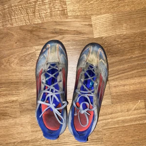 Adidas f50 original elite - Säljer ett par fotbollsskor i blått, rött och silver med snörning och rund tå. Skorna har platt sula med dobbar och är designade för bra grepp på gräs/konstgräs fins original box