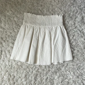 Vit kortkjol med smock från SHEIN - Supersöt vit kortkjol från SHEIN med bred smock i midjan som ger en bekväm och snygg passform. Kjolen har ett lätt utställt fall och är perfekt för varma dagar. Enkel att matcha med olika toppar!