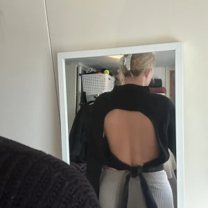 Svart croppad tröja med öppen rygg och knyt - Säljer en svart croppad tröja med öppen rygg och knyt i midjan. Tröjan har lång ärm och är ribbad i materialet, vilket ger en snygg struktur. Perfekt för dig som vill ha något unikt och trendigt!