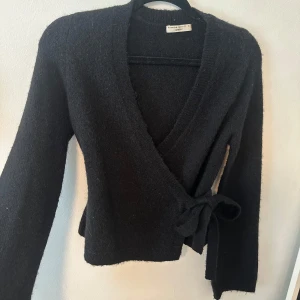 Svart omlottkofta från Gina Tricot - Mysig svart omlottkofta från Gina Tricot med knyt i sidan. Tröjan har lång ärm och är stickad i ett mjukt material. Perfekt att slänga på sig när det är lite kyligare ute. Kommer tolka den innan den säljs, så försvinner små pluttarna!