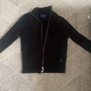 Svart jacka från Tiger of Sweden med dragkedja - Svart jacka från Tiger of Sweden med silverfärgad dragkedja och ribbade muddar. Jackan har ståkrage och två fickor framtill. Enkel och stilren design, perfekt för en clean look. Det är en jacka/zip kofta köpt för 1600kr ungefär. Helt ny använd nån enstaka gång priset kan själv diskuteras
