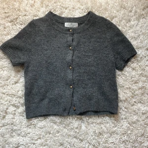 Grå kortärmad kofta med guldiga öppningsbara pärlor från H&M  - Mysig grå kofta från H&M med korta ärmar och guldiga pärlor på framsidan. Koftan är stickad och är mörkgrå. Använd 3-4 gånger. Orginalpris 200kr❣️