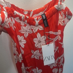 Röd blommig långklänning från ZARA - Säljer en röd långklänning från ZARA med vita blommor och smala axelband. Klänningen har en luftig passform och är perfekt för varma dagar. Materialet känns lätt och bekvämt mot huden. Supersnygg till sommaren!
