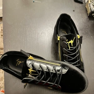 Svarta sneakers från Giuseppe Zanotti med gulddetaljer - Säljer ett par svarta sneakers från Giuseppe Zanotti med guldfärgade dragkedjor och logga. Skorna har snörning, rund tå och är tillverkade i en mix av mocka och skinn. Snygg och lyxig design med svarta sulor och glänsande detaljer.