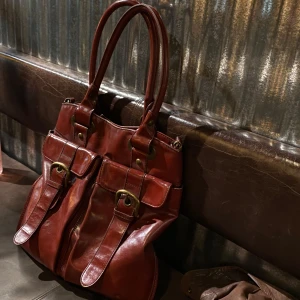 Skitsnygg skinnväska🥰 - Säljer en snygg röd  handväska i skinn med två stora fickor framtill och dekorativa spännen i guld. Väskan har dubbla handtag och rymmer enkelt en laptop. Har även ett långt band som man kan använda! Perfekt accessoar för dig som vill ha både stil och funktion.