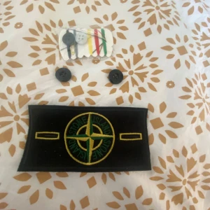 Stone Island tygpatch med knappar - Säljer en klassisk svart och gul Stone Island tygpatch med tillhörande svarta knappar. Perfekt för att sy fast på jacka eller tröja. Märket har broderad logga i grönt och gult.