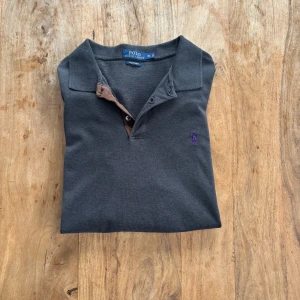 Polo Ralph Lauren – Mörkgrå långärmad piké med lila logga - Klassisk långärmad piké med stretch, kontrastkrage och lila broderad logga. Snygg och ovanlig detalj med färgen!  Storlek: XL (riktigt liten i storleken, skulle säga att den passar M - L)  Färg: Mörkgrå med brun krage och lila logga Material: Stretch Mesh (bomull med stretch) Skick: Mycket fint skick – inga fläckar eller hål Stil: Sportig / Avslappnad / Vintage edge Passform: Klassisk rak modell Nypris: ca 1 400 kr Pris: (300 kr)  Frakt: Skickas snabbt & spårbart