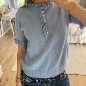 Ljusblå stickad topp med volang och pärlknappar från Studio 21 - Supersöt ljusblå stickad topp från Studio 21 med korta ärmar och volangdetaljer vid hals och ärmslut, dekorerad med glittriga pärlknappar. Perfekt för dig som gillar romantiska detaljer och mjuka material!🤩