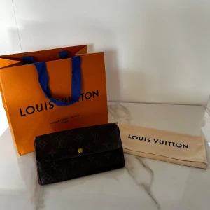 Louis Vuitton plånbok i monogram canvas - Säljer en klassisk Louis Vuitton plånbok i mörkbrun monogram canvas med guldknapp. Insidan är brun med dragkedjefack och flera fack för kort och sedlar. Kommer med dustbag och originalpåse. Plånboken är i acceptabelt skick men har defekter på sidorna, därav det låga priset. Plånboken har blivit autentiserad av Tradera.