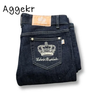 Bootcut Victoria Beckham Jeans - Säljer ett par mörkblå jeans med snygg broderad krona och text på bakfickorna. Jeansen har klassisk femficksdesign, raka ben och läderpatch bak i midjan. Perfekta för dig som vill sticka ut lite extra med detaljer.