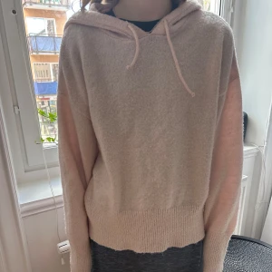 Ljusrosa stickad hoodie - Mysig ljusrosa stickad hoodie med huva. Tröjan har långa ärmar och ribbade muddar vid ärmslut och nederkant. Perfekt för kyliga dagar när du vill vara både bekväm och snygg.