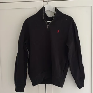 Svart halvzip tröja från Polo Ralph Lauren - Svart långärmad halvzip tröja från Polo Ralph Lauren med röd broderad logga på bröstet. Klassisk och stilren modell med hög krage och ribbade muddar. Perfekt för en avslappnad men snygg look.