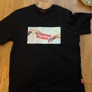 Svart Supreme t-shirt med konstmotiv - Säljer en svart t-shirt från Supreme med ett konstinspirerat tryck på bröstet där två händer möts över Supreme-loggan. T-shirten är i 100% bomull och har normal passform. Perfekt för dig som gillar streetwear och snygga prints.