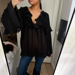 Svart blus med volangdetaljer - Säljer en svart blus med v-ringning och stora volangdetaljer längs halsringningen och ärmarna. Blusen har långa, vida ärmar och är tillverkad i ett lätt, transparent material.