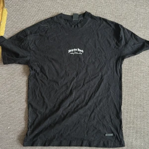 Svart t-shirt med tryck 'Drip Too Hard' - Svart t-shirt med vitt tryck på bröstet och stort tryck på ryggen med texten 'Drip Too Hard'. Klassisk passform och rund halsringning. Perfekt för en streetwear-look.