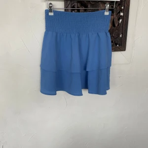 Blå volangkjol från ONLY - Säljer en blå kortkjol från ONLY med smock i midjan och dubbla volanger. Kjolen är luftig och har en fin struktur i tyget, perfekt för varma dagar. Enkel att matcha med olika toppar för en trendig look.