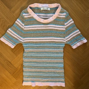 Randig topp från Design by Si - Superfin topp med korta ärmar från Design by Si. Tröjan har randigt mönster i pastellrosa, blått och beige med glittriga inslag. Mjuk ribbad kant vid hals, ärmslut och nederkant. Perfekt för vår och sommar!