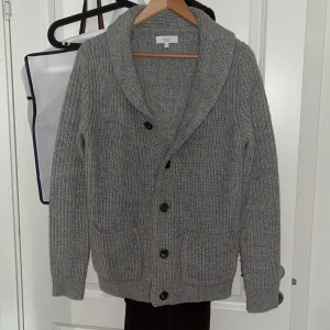 Grå stickad kofta från Next - Mysig grå stickad kofta från Next med stora knappar framtill och två fickor. Koftan har en bred krage och är perfekt för lager-på-lager. Passformen är normal och materialet känns mjukt och värmande. Perfekt under sommarkvällar. Storlek S, använd fåtal gånger (skick som nytt) alltså inga defekter osv.