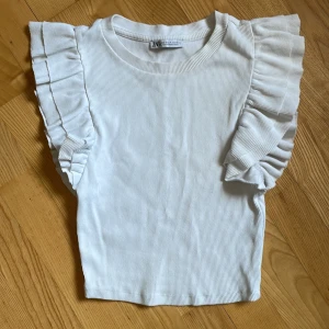 Vit ribbad topp med volangärmar från Zara - Säljer en söt vit ribbad topp från Zara med volangdetaljer längs ärmarna. 30 kr.