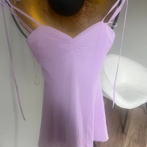 Ljuslila axelbandslös klänning - Söt ljuslila klänning med smala axelband och lätt rynkning vid bysten. Klänningen har en feminin siluett och är perfekt för varma dagar. Materialet känns lätt och luftigt, och modellen är enkel men stilren. Den har även knytning därbak 