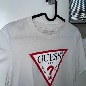 Vit t-shirt från Guess med logga - Vit t-shirt från Guess med den klassiska röda triangelloggan framtill. Modellen är slim fit och tillverkad i ekologisk bomull. Perfekt för en clean och stilren look.