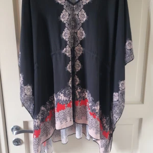 Svart och mönstrad klänning från Bodyflirt - Säljer en svart klänning från Bodyflirt med vackert vitt och rött mönster längs kanterna och nertill. Klänningen har lös passform och är perfekt för dig som gillar bohemisk stil. Den har vida ärmar och ett snyggt fall.