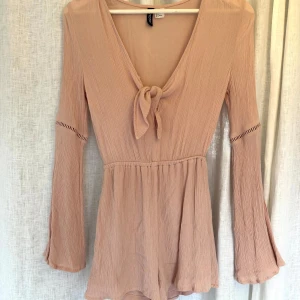 Rosa jumpsuit  - Säljer en rosa jumpsuit med lång ärm och knyt framtill. Jumpsuiten har v-ringning och är tillverkad i ett lätt, strukturerat tyg som ger en luftig känsla. Perfekt för vår och sommar! Hade själv denna på min studentmottagning och passar både till fest och vardag! 