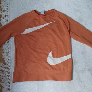 Orange sweatshirt från Nike med stora vita swoosh-loggor - Säljer en orange sweatshirt från Nike med stora vita swoosh-loggor både fram och bak. Tröjan har rund halsringning och långa ärmar. Perfekt för en sportig och avslappnad stil.
